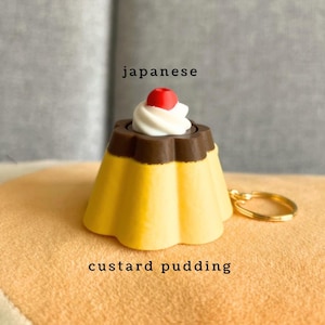 May include: A keyring featuring a miniature custard pudding charm with a yellow base, a brown top layer, a white whipped cream swirl, and a small red cherry on top. The text "japanese" and "custard pudding" is printed above and below the charm.