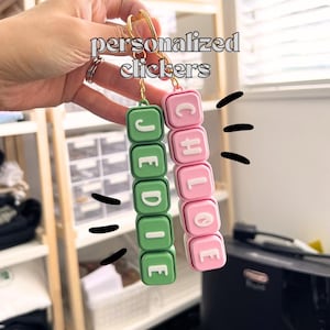 May include: Two personalised clickers, one green and one pink, spelling out "JEDIE" and "CHLOE" in white letters. The clickers are attached to gold keychains. The image has the text "personalised clickers" at the top.