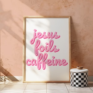 K&ouml;nnte beinhalten: Ein gerahmter Kunstdruck mit dem Schriftzug "jesus foils caffeine" in rosa. Der Druck befindet sich in einem goldenen Rahmen und lehnt an einer pfirsichfarbenen Wand. Daneben steht ein schwarz-wei&szlig; karierter Beh&auml;lter mit Holzdeckel.