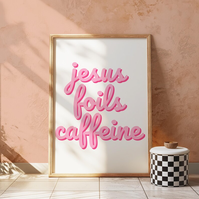 K&ouml;nnte beinhalten: Ein gerahmter Kunstdruck mit dem Schriftzug "jesus foils caffeine" in rosa. Der Druck befindet sich in einem goldenen Rahmen und lehnt an einer pfirsichfarbenen Wand. Daneben steht ein schwarz-wei&szlig; karierter Beh&auml;lter mit Holzdeckel.