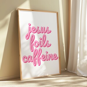 K&ouml;nnte beinhalten: Ein gerahmter Kunstdruck mit dem Schriftzug "jesus foils caffeine" in rosa Schrift. Die W&ouml;rter haben einen Schatteneffekt, und der Druck befindet sich in einem hellen Holzrahmen. Der Hintergrund ist wei&szlig;.