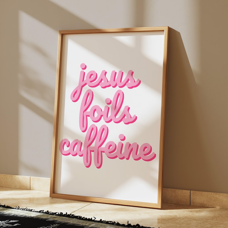 K&ouml;nnte beinhalten: Gerahmter Druck mit den Worten "jesus foils caffeine" in rosa, kursiver Schrift. Der Druck hat einen wei&szlig;en Hintergrund und einen hellbraunen Holzrahmen. Das Kunstwerk lehnt an einer beigen Wand.