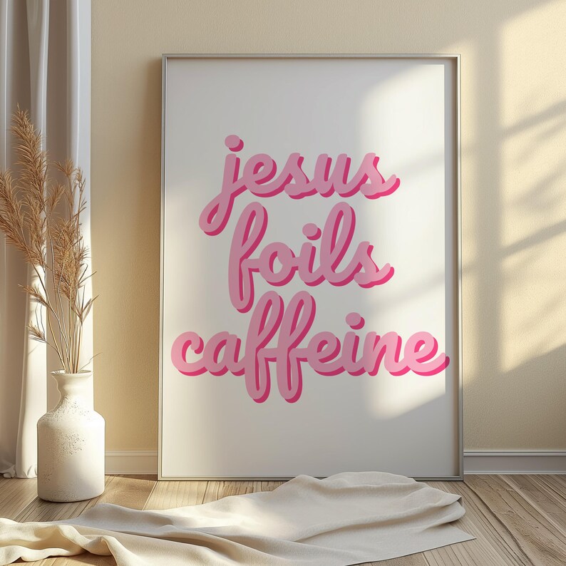 K&ouml;nnte beinhalten: Gerahmter Kunstdruck mit den Worten "Jesus foils caffeine" in rosa, kursiver Schrift. Das Kunstwerk ist vor einem wei&szlig;en Hintergrund platziert und in einem silbernen Rahmen gerahmt. Der Druck wird auf einem Holzboden ausgestellt.