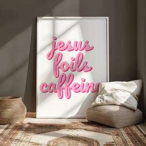 K&ouml;nnte beinhalten: Ein gerahmter Kunstdruck mit den Worten "Jesus foils caffeine" in rosa, stilisierten Buchstaben. Der Druck wird vor einer neutralen Wand ausgestellt, mit einem geflochtenen Korb und einem Bodenkissen im Vordergrund. Der Raum hat einen Teppich mit einem braunen und beigen Muster.