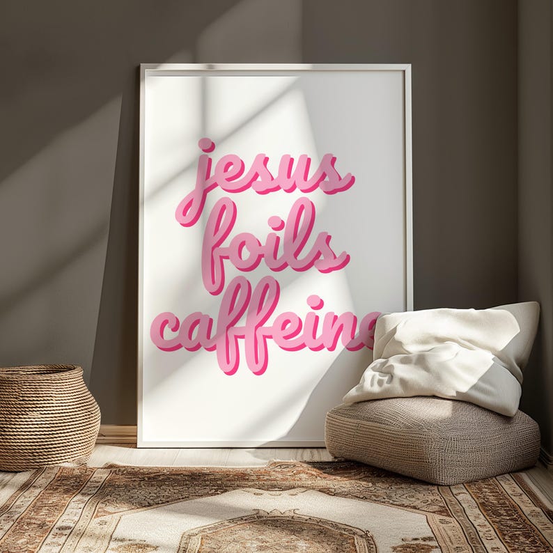 K&ouml;nnte beinhalten: Ein gerahmter Kunstdruck mit den Worten "Jesus foils caffeine" in rosa, stilisierten Buchstaben. Der Druck wird vor einer neutralen Wand ausgestellt, mit einem geflochtenen Korb und einem Bodenkissen im Vordergrund. Der Raum hat einen Teppich mit einem braunen und beigen Muster.