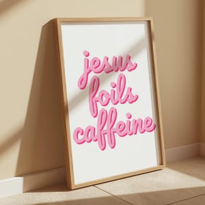 K&ouml;nnte beinhalten: Ein gerahmter Kunstdruck mit dem Schriftzug "Jesus foils caffeine" in rosa Schrift auf wei&szlig;em Hintergrund. Der Holzrahmen lehnt an einer Wand. Der Text hat einen 3D-Effekt mit einem dunkleren rosa Schatten.