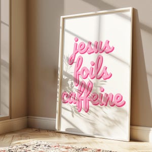 K&ouml;nnte beinhalten: Gerahmter Kunstdruck mit dem Schriftzug "Jesus Foils Caffeine" in rosa Schrift auf cremefarbenem Hintergrund. Das Kunstwerk befindet sich in einem wei&szlig;en Rahmen. Das Bild ist gut beleuchtet mit Schatten.