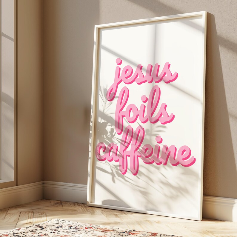 K&ouml;nnte beinhalten: Gerahmter Kunstdruck mit dem Schriftzug "Jesus Foils Caffeine" in rosa Schrift auf cremefarbenem Hintergrund. Das Kunstwerk befindet sich in einem wei&szlig;en Rahmen. Das Bild ist gut beleuchtet mit Schatten.