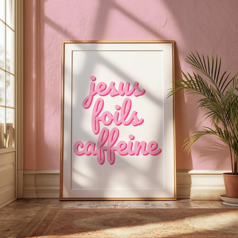 K&ouml;nnte beinhalten: Gerahmter Kunstdruck mit dem Schriftzug "jesus foils caffeine" in rosa Schrift. Das Kunstwerk in Goldrahmen ist vor einer rosa Wand platziert. Rechts steht eine Topfpflanze. Sonnenlicht f&auml;llt durch ein Fenster.