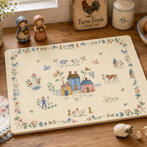 May include: A rectangular cream-colored cutting board featuring a central illustration of three houses surrounded by farm animals, flowers, and a person gardening. The border is decorated with a repeating pattern of small flowers and birds.