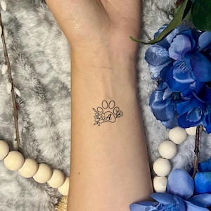 May include: A black paw print tattoo with floral accents and the letter "A" on a person's wrist. The arm is surrounded by blue flowers, white wooden beads, and decorative branches.