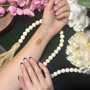 May include: A red, anatomical heart temporary tattoo is displayed on a person's wrist. The image is surrounded by white beads, pink flowers, and a white peony. The person's nails are painted with a black French tip.