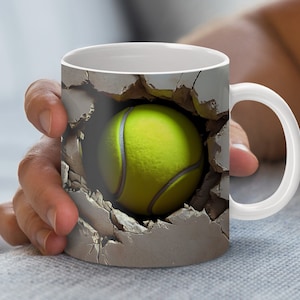 May include: White ceramic coffee mug featuring a tennis ball design. The tennis ball appears to be breaking through a wall, with a realistic texture and color. The mug has a white handle and a smooth, glossy finish.