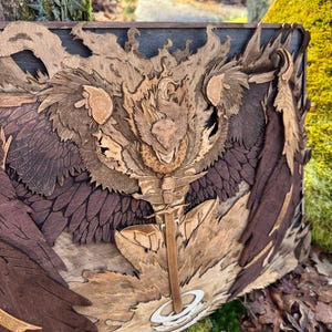 Puede incluir: Una pieza de arte de pared de madera que representa una criatura alada con un halo de fuego. La criatura está tallada en detalle y tiene una espada en la mano. La obra de arte está hecha de diferentes tonos de madera, incluyendo marrón, rojo y dorado.