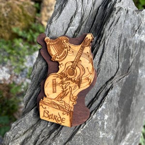 May include: A wooden cutout of a bard playing a guitar and a harp. The cutout is brown and has the word "Barde" written on it.