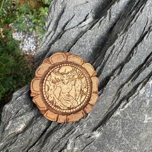 May include: A wooden round coaster with a detailed wood-burned design of a woman with wings and a flowing dress. The coaster has a decorative border with a repeating pattern.