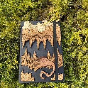 Peut inclure: Un carnet noir avec une couverture en bois présentant un motif découpé au laser d'une chaîne de montagnes et d'une créature mythique. Le carnet est doté d'une fermeture à élastique noire.
