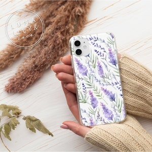 Op de afbeelding: Een transparante telefoonhoes met een aquarelontwerp van lavendelbloemen en groene bladeren op een witte achtergrond. De hoes wordt vastgehouden door een hand die een beige trui draagt.