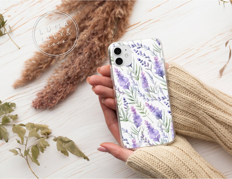Op de afbeelding: Een transparante telefoonhoes met een aquarelontwerp van lavendelbloemen en groene bladeren op een witte achtergrond. De hoes wordt vastgehouden door een hand die een beige trui draagt.