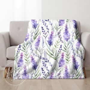Op de afbeelding: Een witte deken met een lavendelbloemenpatroon. De deken heeft aquarel-achtige paarse en groene lavendelbloemen op een witte achtergrond.