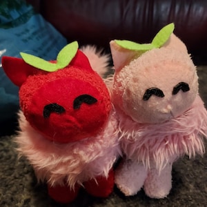 以下が含まれることがあります： 緑のフェルトの葉っぱが頭に乗った、赤い猫とピンクの猫のぬいぐるみ2つ。どちらも黒い目とピンクの毛並みです。