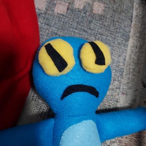 May include: A blue plush toy with yellow eyes and a black frown. The toy has a light blue belly and outstretched arms.