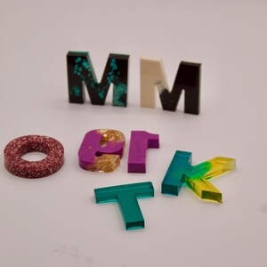 May include: A set of colourful resin letters, including the letters M, O, T, K, and 1. The letters are made of clear resin with different colours and patterns, including green, red, purple, and gold.