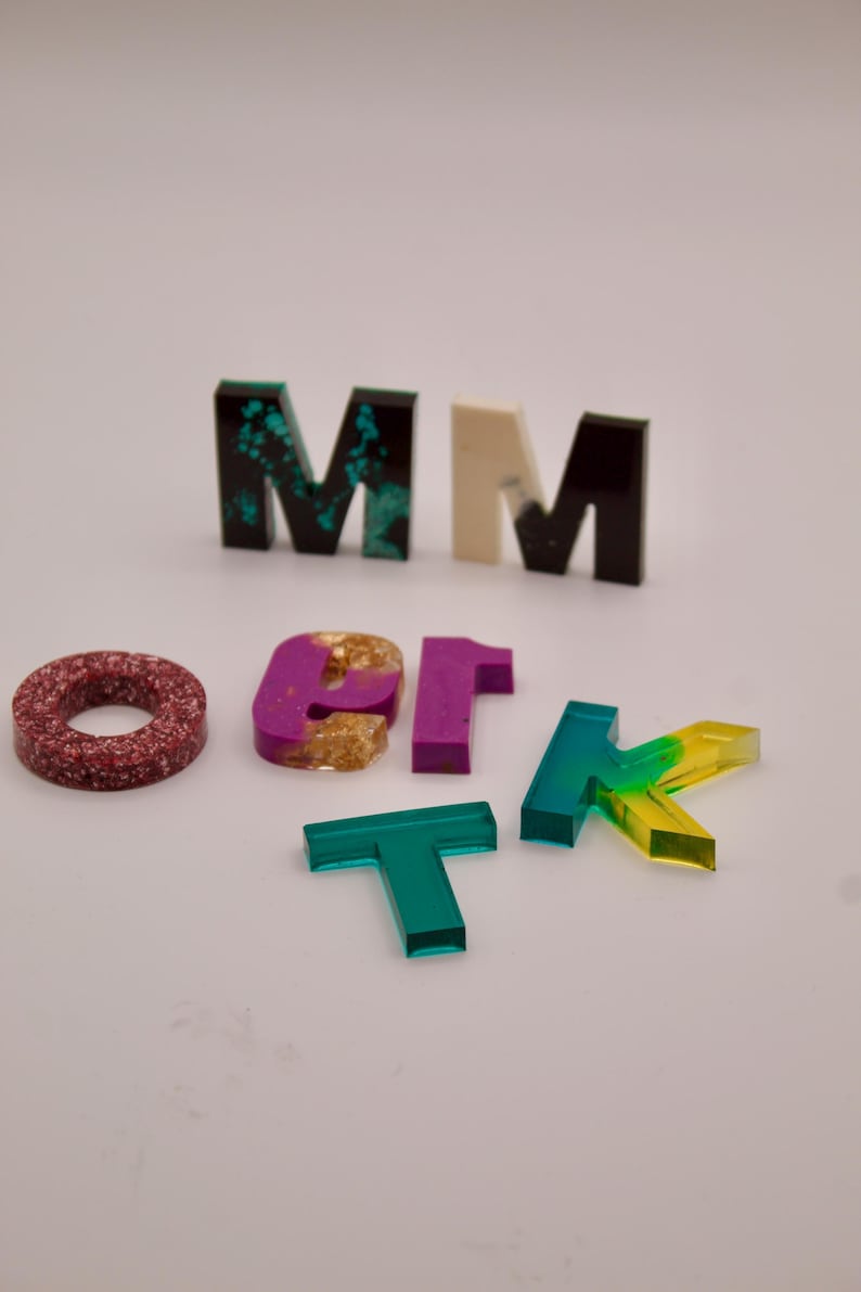May include: A set of colourful resin letters, including the letters M, O, T, K, and 1. The letters are made of clear resin with different colours and patterns, including green, red, purple, and gold.