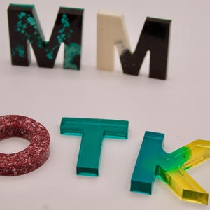 May include: A set of colourful resin letters spelling out "MMOTK". The letters are made of clear resin with different colours and patterns. The "M" is green with black and white flecks, the second "M" is white with black flecks, the "O" is red with white flecks, the "T" is green, and the "K" is green and yellow.