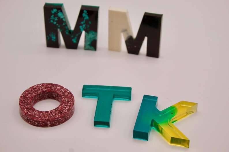 May include: A set of colourful resin letters spelling out "MMOTK". The letters are made of clear resin with different colours and patterns. The "M" is green with black and white flecks, the second "M" is white with black flecks, the "O" is red with white flecks, the "T" is green, and the "K" is green and yellow.