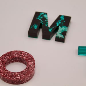 May include: Close-up of three letters, M, O, and T, made of resin. The M and T are black with turquoise accents. The O is red with white flecks.