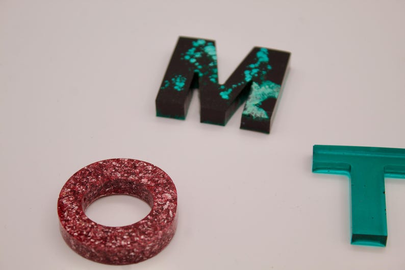 May include: Close-up of three letters, M, O, and T, made of resin. The M and T are black with turquoise accents. The O is red with white flecks.