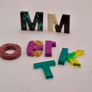 May include: A set of colourful resin letters, including a green "M", a white "M", a black "M", a red circle, a purple "1", a gold "O", a green "T", and a yellow and green "K".