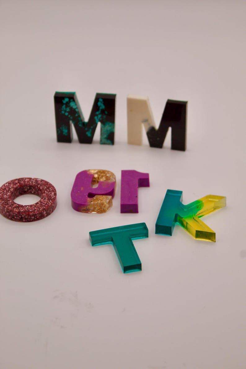 May include: A set of colourful resin letters, including a green "M", a white "M", a black "M", a red circle, a purple "1", a gold "O", a green "T", and a yellow and green "K".