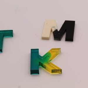 May include: A set of colourful acrylic letters, including a green "T", a yellow and green "K", a white and black "M", and a black "M".