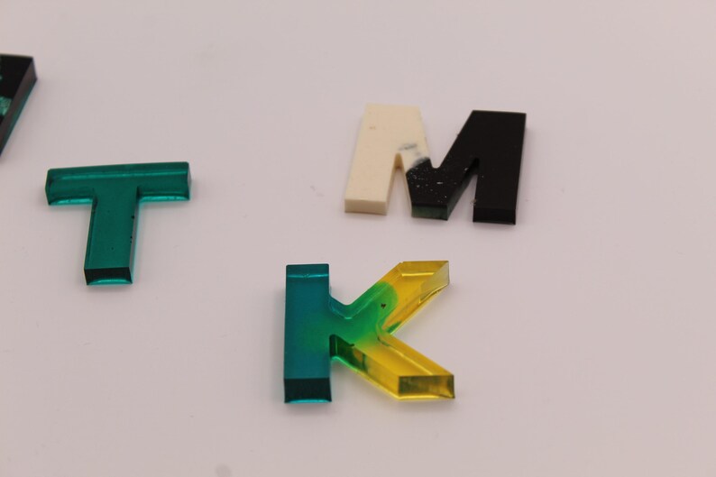 May include: A set of colourful acrylic letters, including a green "T", a yellow and green "K", a white and black "M", and a black "M".