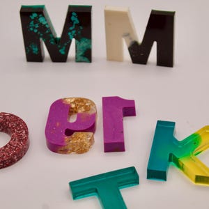 May include: A set of resin letters in various colours and styles, including a red glitter "O", a purple and gold "er", a green and yellow "K", a teal "T", and two "M"s in black, white, and teal.