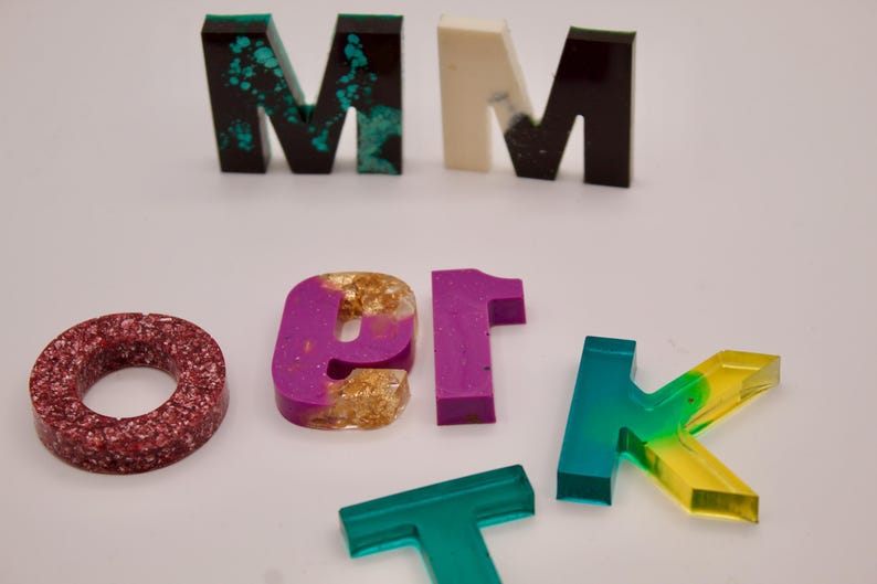 May include: A set of resin letters in various colours and styles, including a red glitter "O", a purple and gold "er", a green and yellow "K", a teal "T", and two "M"s in black, white, and teal.
