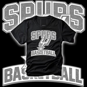May include: Black t-shirt with the text "SPURS BASKETBALL" in white and gray. The shirt features the Spurs logo, a stylized "SPURS" wordmark, and a basketball graphic. The design is set against a black background.