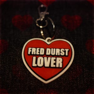 May include: A heart-shaped keychain tag with the words "FRED DURST LOVER" in white block letters on a red background. The tag has a white border and a silver-colored clasp and ring.