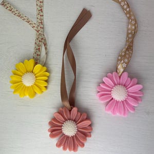 May include: Three daisy-shaped ornaments in yellow, pink, and coral, each with a white center. They hang from decorative ribbons in various patterns and colors, perfect for adding a touch of spring to any space.