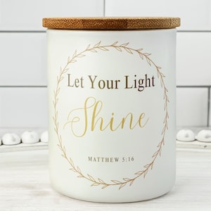 May include: A white candle with a wooden lid, featuring the text "Let Your Light Shine" in gold script, surrounded by a gold wreath. The text "Matthew 5:16" is below. The candle sits on a white tray.