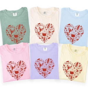 Könnte beinhalten: Sechs farbige T-Shirts in Pastelltönen von Grün, Creme, Blau, Rosa, Lila und Gelb. Jedes Shirt hat ein rotes herzförmiges Design mit den Worten "Mudbug Cajun Pinch Peel" und Illustrationen von Krebsen, Mais und einem Topf.
