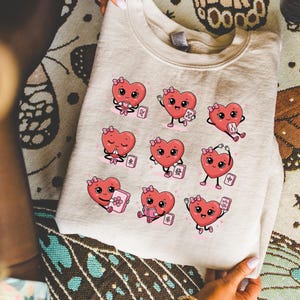 Peut inclure: Un sweat-shirt beige à col rond orné d'un imprimé graphique représentant neuf cœurs de dessin animé dans diverses poses, chacun interagissant avec une tuile de Mahjong. Les cœurs sont roses avec de petits nœuds et les tuiles affichent des symboles.