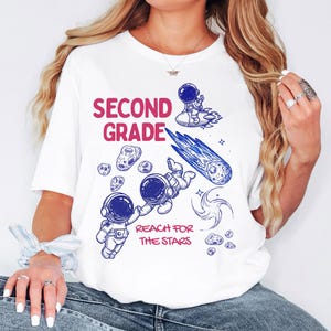 Puede incluir: Una camiseta blanca con un estampado gráfico que incluye el texto "SECOND GRADE" y "REACH FOR THE STARS" en rosa. El diseño presenta ilustraciones en línea azul de dos astronautas flotando en el espacio, un cohete, un cometa y asteroides.
