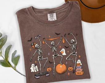 Könnte beinhalten: Ein braunes T-Shirt mit einer Grafik von drei Skeletten, die mit einer Hantel, einem Widerstandsband und einem Gymnastikball trainieren. Das Design enthält kleine Geister, Fledermäuse, Kürbisse und Blöcke mit dem Text MOVE PLAY GROW THRIVE.