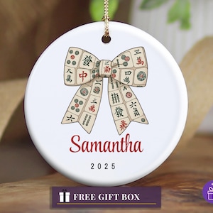 Könnte beinhalten: Runder weißer Keramikschmuck mit einer goldenen Aufhängeschnur. Er zeigt ein Schleifendesign aus Mahjong-Steinen. Der Name "Samantha" ist rot aufgedruckt, darunter das Jahr "2025". Unten befindet sich ein Geschenkbox-Symbol.