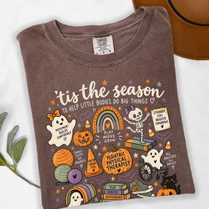 May include: A brown short-sleeve t-shirt featuring the text 'tis the season to help little bodies do big things' and graphic illustrations of ghosts, pumpkins, a rainbow, a skeleton, a wheelchair, and books with various inspirational phrases.
