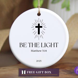 Puede incluir: Adorno navideño redondo de cerámica blanca con un diseño de cruz negra y las palabras "BE THE LIGHT" y "Matthew 5:14". El año "2025" está impreso debajo. Una cuerda dorada está adjunta.
