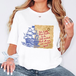 May include: White t-shirt featuring a blue illustration of a sailing ship on the ocean. The design includes a parchment with the quote "We hold these truths to be self evident that all men are created equal."